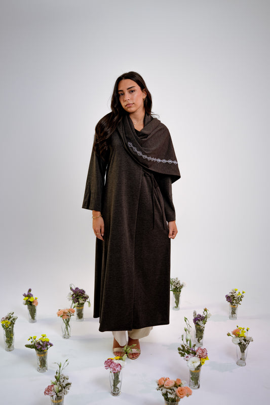 Woman wearing a dark gray abaya with n embroidered shawl standing in the middle of small floral arrangements on a light background