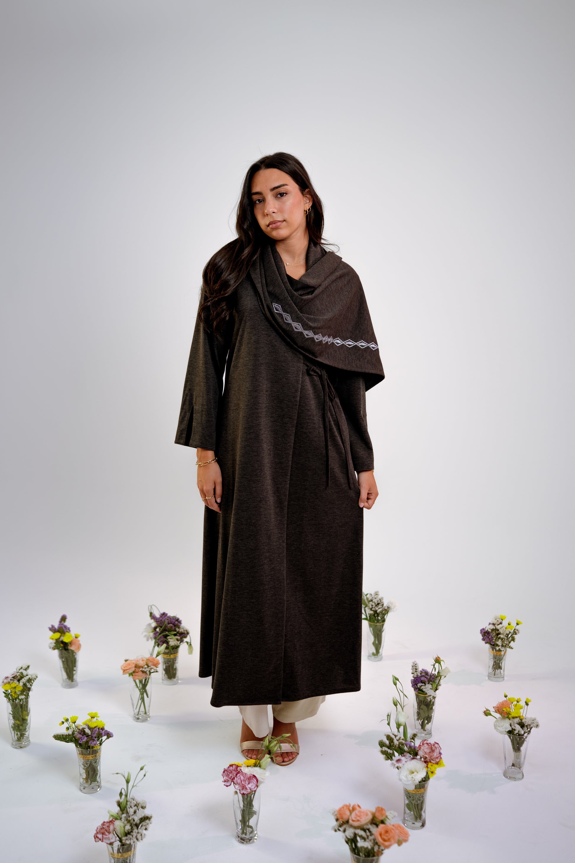 Woman wearing a dark gray abaya with n embroidered shawl standing in the middle of small floral arrangements on a light background
