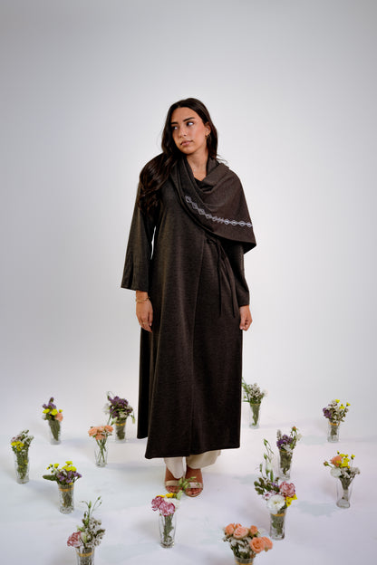 Woman wearing a dark gray abaya with n embroidered shawl standing in the middle of small floral arrangements on a light background