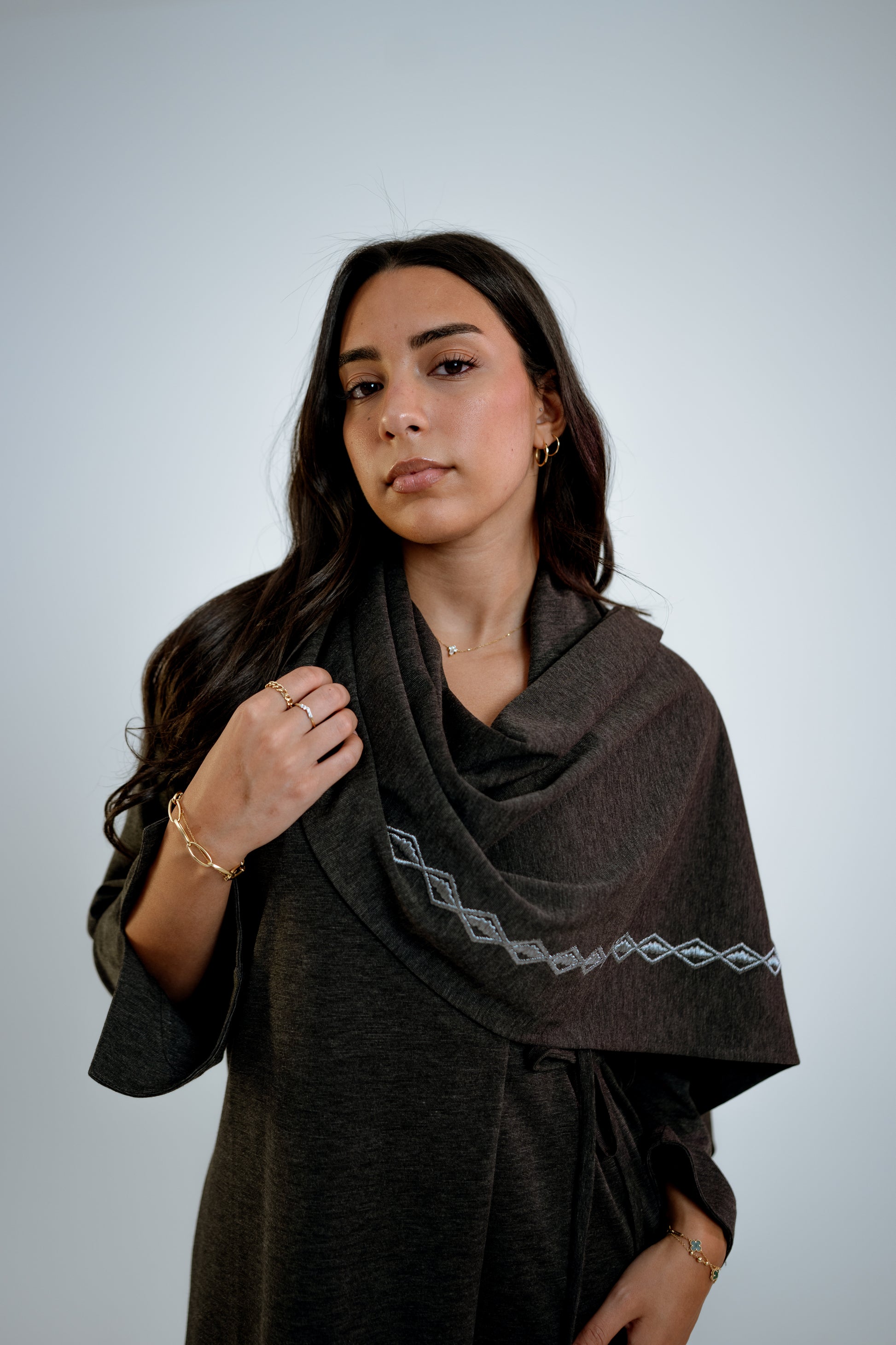 Woman wearing a dark gray abaya with n embroidered shawl standing in front of a light background