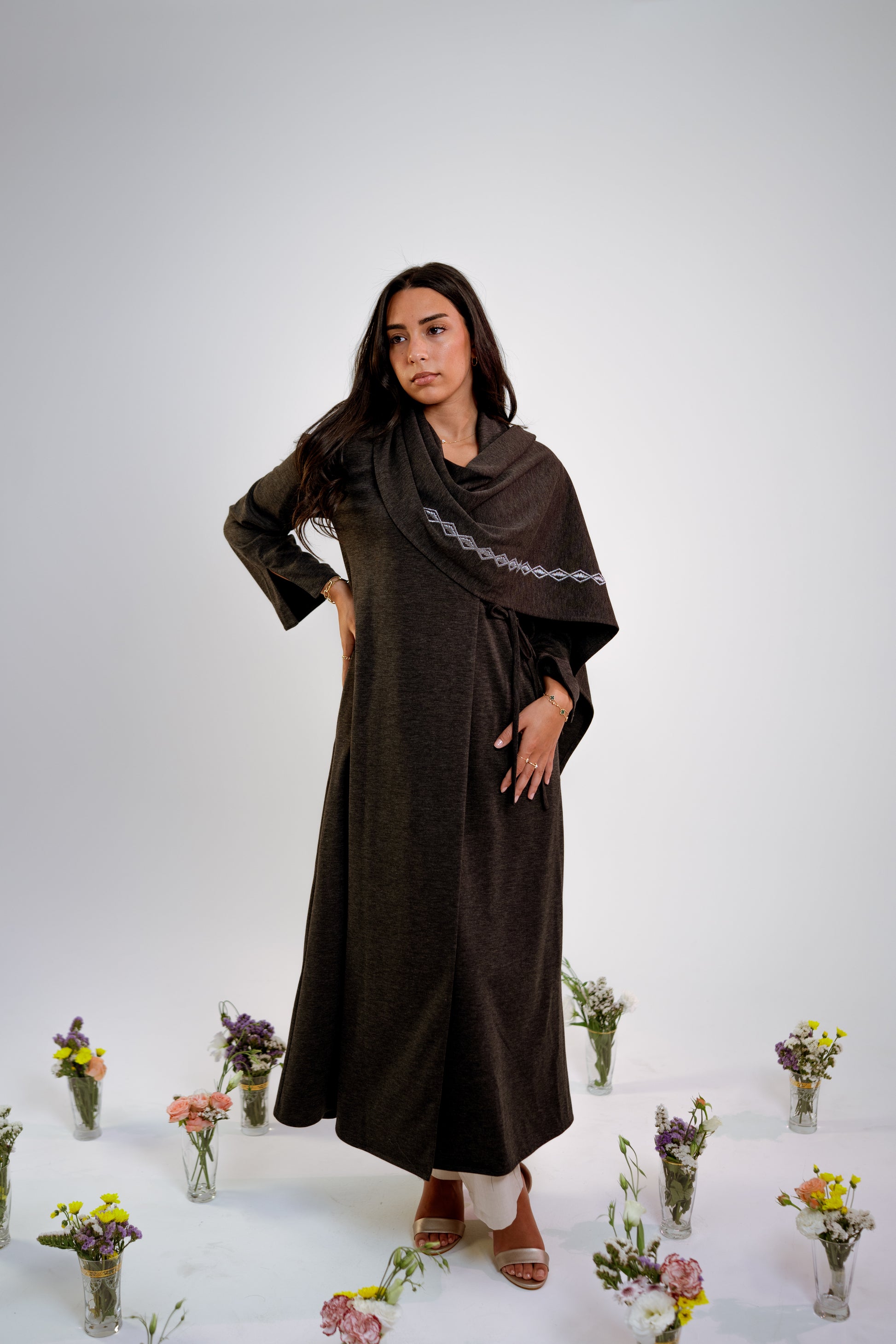 Woman wearing a dark gray abaya with n embroidered shawl standing in the middle of small floral arrangements on a light background
