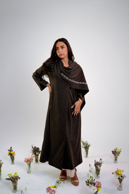 Woman wearing a dark gray abaya with n embroidered shawl standing in the middle of small floral arrangements on a light background