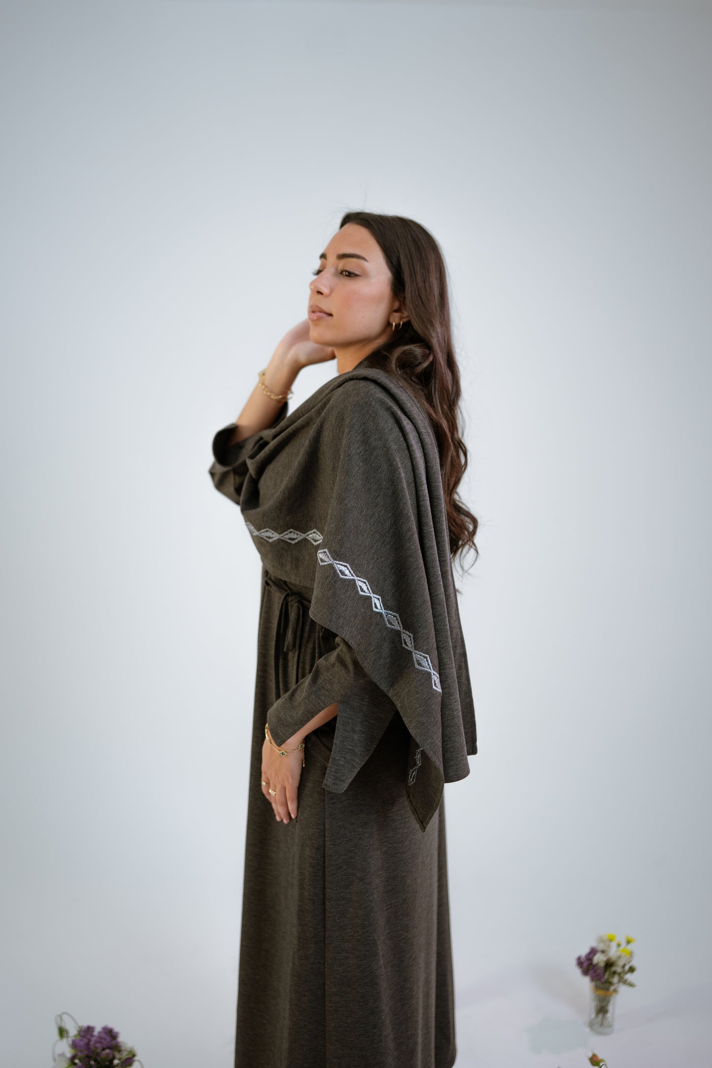 Woman wearing a dark gray abaya with n embroidered shawl standing sideways in the middle of small floral arrangements on a light background