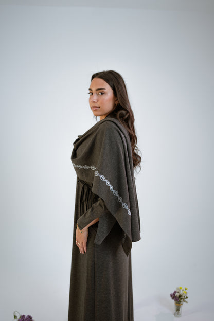 Woman wearing a dark gray abaya with n embroidered shawl standing sideways in the middle of small floral arrangements on a light background