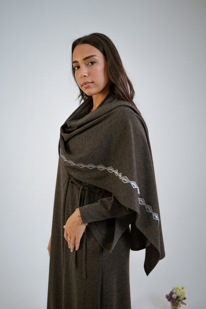 Woman wearing a dark gray abaya with n embroidered shawl standing sideways in the middle of small floral arrangements on a light background