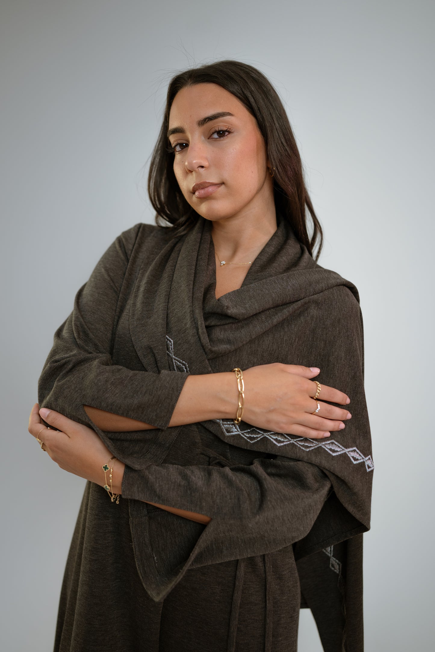 Woman wearing a dark gray abaya with n embroidered shawl standing in front of a light background