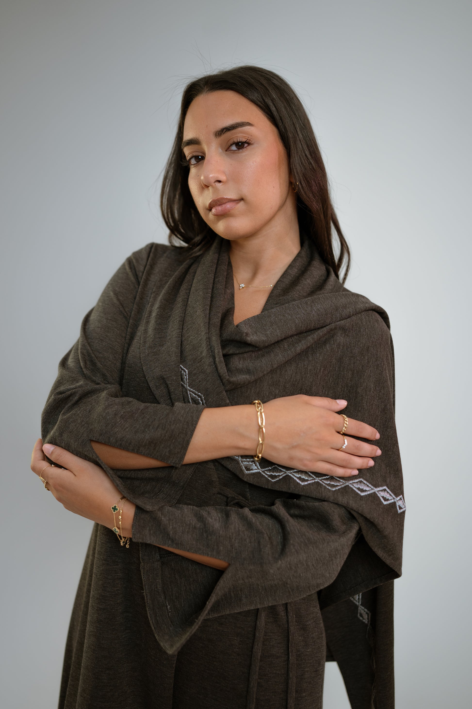 Woman wearing a dark gray abaya with n embroidered shawl standing in front of a light background